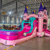 Side view of Grand Royal Castle Commercial Econo Inflatable Combo (Wet/Dry) set up in a large indoor warehouse. Features pink castle structure with purple turrets, yellow flags, teal slide, and pink splash pool. Metal roofing, disco ball, and other warehouse equipment visible, showcasing its size in a commercial setting.