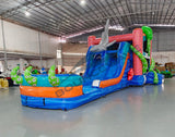 Front view of Hammerhead Reef Dual Lane Commercial Inflatable Combo featuring a vibrant ocean-themed design with dual blue slides, a large green octopus on top, and inflatable shark fins. The structure includes a pink coral-patterned bounce house area, mesh windows, and a splash pool. Set up in a spacious warehouse with metal roofing and other inflatable products visible in the background.