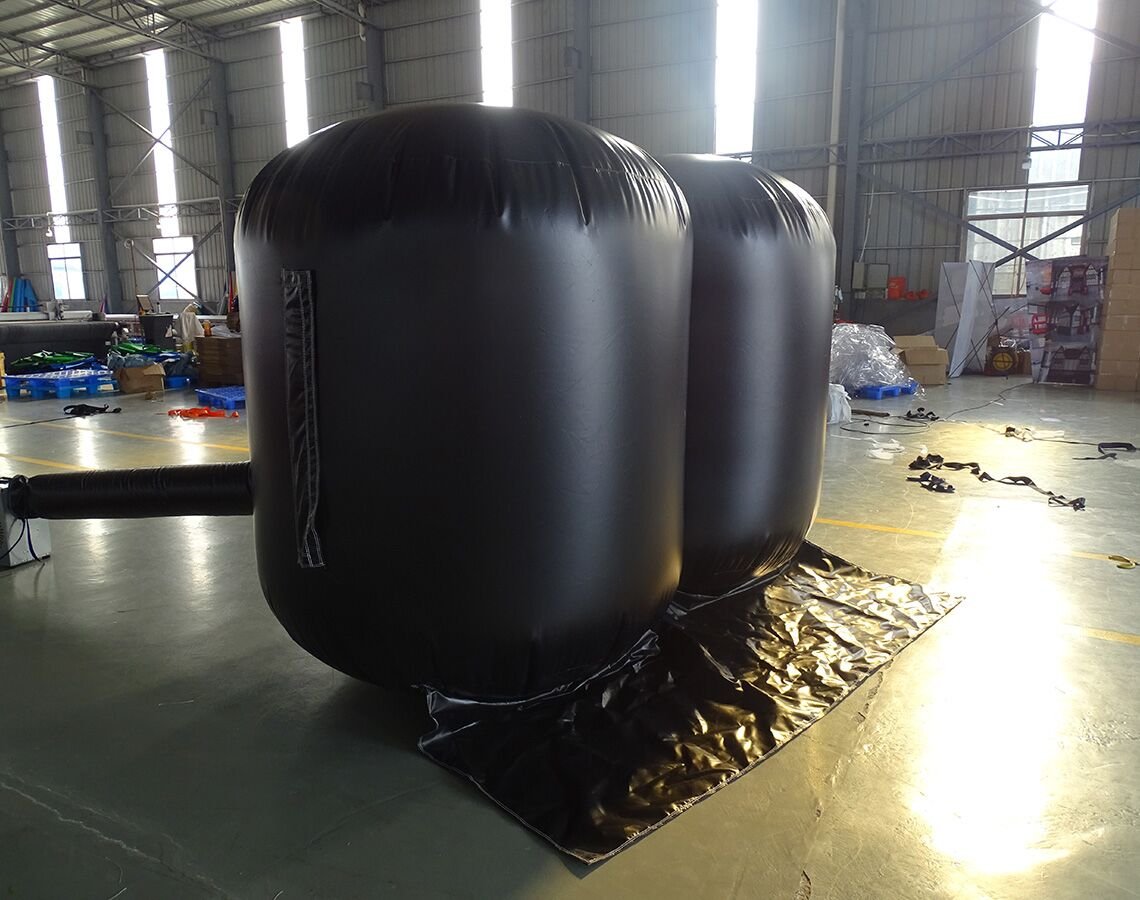 Side view of two large black Inflatable Lifting Wedges in a warehouse setting. The wedges are fully inflated, showing their cylindrical shape. Bright natural light streams in from windows, illuminating the industrial space with various equipment and materials visible in the background.
