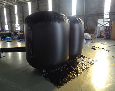 Side view of two large black Inflatable Lifting Wedges in a warehouse setting. The wedges are fully inflated, showing their cylindrical shape. Bright natural light streams in from windows, illuminating the industrial space with various equipment and materials visible in the background.
