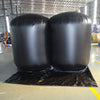Front view of two fully inflated black Inflatable Lifting Wedges in a warehouse. The wedges are positioned side by side on a black protective mat. The industrial setting shows a concrete floor, metal roof structure, and various colorful equipment and materials in the background.