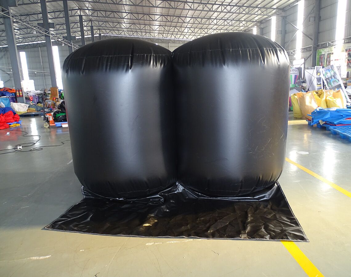 Front view of two fully inflated black Inflatable Lifting Wedges in a warehouse. The wedges are positioned side by side on a black protective mat. The industrial setting shows a concrete floor, metal roof structure, and various colorful equipment and materials in the background.