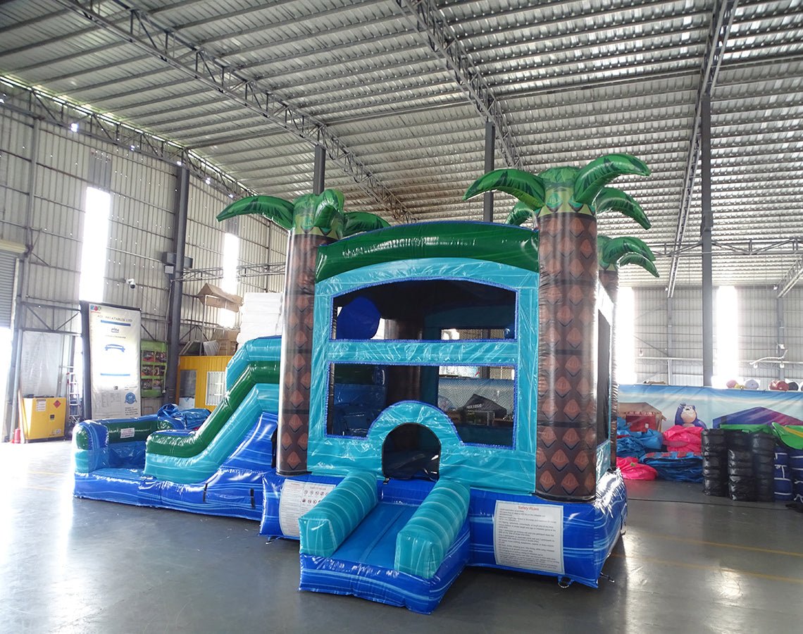 Side angle view of the Island Drop Econo Combo featuring a blue and green bounce house with palm tree decorations, curved slide, and spacious splash pool. The inflatable is set up in an indoor warehouse with metal roofing and other equipment visible.