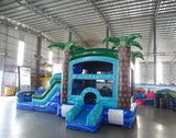 Side angle view of the Island Drop Econo Combo featuring a blue and green bounce house with palm tree decorations, curved slide, and spacious splash pool. The inflatable is set up in an indoor warehouse with metal roofing and other equipment visible.
