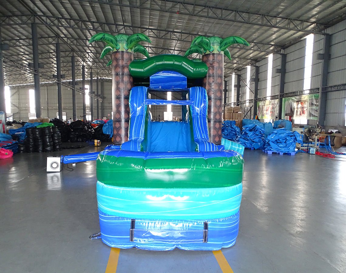 Front view of Island Drop Econo Combo, showcasing the bounce house entrance with palm tree decorations, blue slide, and green splash pool area. Set up in an indoor warehouse with other inflatable products and equipment visible in the background.