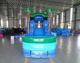 Front view of Island Drop Econo Combo, showcasing the bounce house entrance with palm tree decorations, blue slide, and green splash pool area. Set up in an indoor warehouse with other inflatable products and equipment visible in the background.
