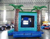 Large inflatable tropical-themed combo featuring a bounce house with palm tree decorations, attached slide, and splash pool. The structure has vibrant blue and green colors with brown palm tree trunks. Set up in an indoor warehouse with metal roofing and other inflatable products visible in the background.