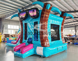 Front view of Island Flow Econo Combo, showcasing the bounce house entrance with palm tree decorations, colorful slide, and splash pool area. Set up in an indoor warehouse with metal roofing and other inflatable products visible in the background.
