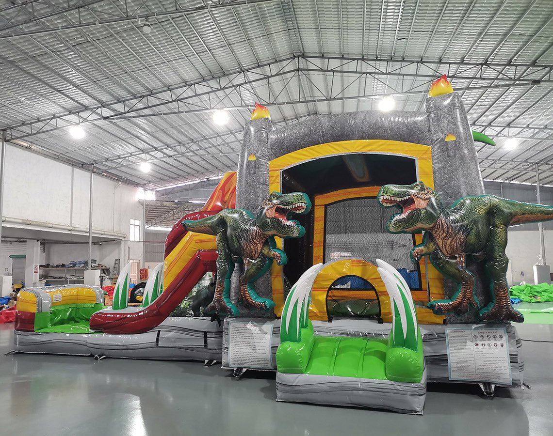 Front view of Lava Raptor Econo Combo, showcasing the volcano entrance with lava design, red slide, and green splash pool area. Large, lifelike dinosaur figures flank the entrance. Displayed in an indoor warehouse with metal roofing and other equipment visible.