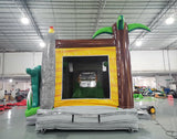 Large inflatable dinosaur-themed combo featuring a volcano-shaped bounce house with lava design, attached slide, and splash pool. The structure has vibrant red, orange, and green colors with realistic T-Rex dinosaur figures guarding the entrance. Set up in an indoor warehouse with metal roofing and other inflatable products visible in the background.