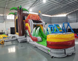 Side view of Lava Raptor Econo Combo showing the volcano-shaped bounce house, red slide, and splash pool area. Features bright red, orange, and green colors with palm tree decorations and dinosaur figures. Set up in an indoor warehouse with other inflatable products visible.