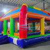 Side view of Mega Fun Dome Commercial Grade Bounce House set up in a large indoor warehouse. The inflatable features a rounded top with blue, yellow, green, and red panels. Green support pillars and orange beams frame the structure. Mesh windows provide visibility and ventilation. The warehouse setting shows metal roofing and other inflatable products in the background.