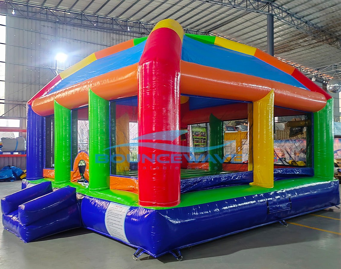 Side view of Mega Fun Dome Commercial Grade Bounce House set up in a large indoor warehouse. The inflatable features a rounded top with blue, yellow, green, and red panels. Green support pillars and orange beams frame the structure. Mesh windows provide visibility and ventilation. The warehouse setting shows metal roofing and other inflatable products in the background.