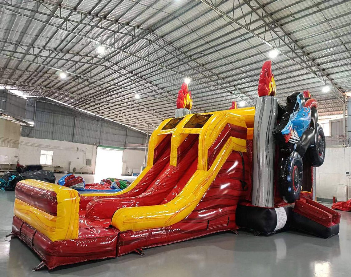 Cross Front view of Monster Truck Dual Lane Commercial Inflatable Combo (Wet/Dry) set up in a large indoor warehouse. The inflatable features dual slides in red and yellow, leading to a splash pool area. A blue monster truck with flame decals is mounted on top of gray pillars with flame decorations. The base is predominantly red with yellow accents. Various other inflatable products and equipment are visible in the background, showcasing the warehouse setting with metal roofing and bright lighting.