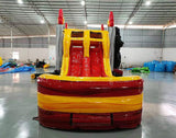 Front view of Monster Truck Dual Lane Commercial Inflatable Combo showing dual slides in red and yellow leading to a splash pool. The structure features a blue monster truck with flame decals mounted on top and flame decorations on the sides. Set up in a warehouse with concrete flooring and various other inflatable products visible in the background.