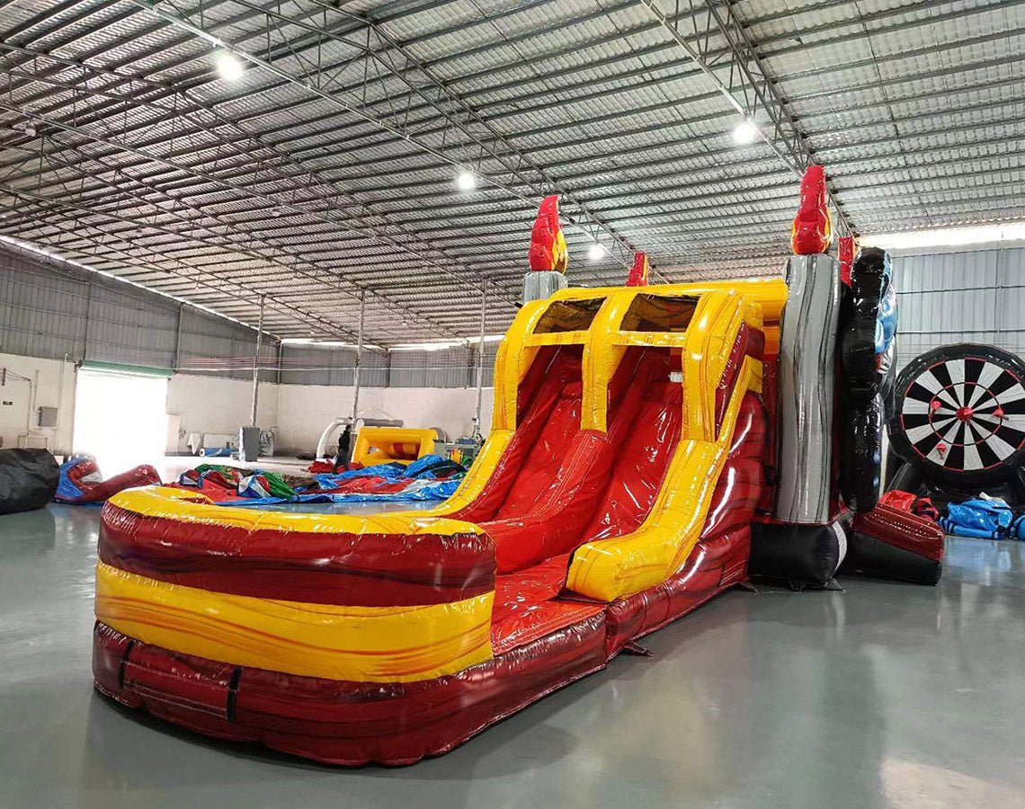 Three-quarter view of Monster Truck Dual Lane Commercial Inflatable Combo in a spacious warehouse. The inflatable displays a blue monster truck on top, dual slides in red and yellow, and a large splash pool area. Other inflatable products and warehouse materials are visible in the background, showcasing its commercial application.