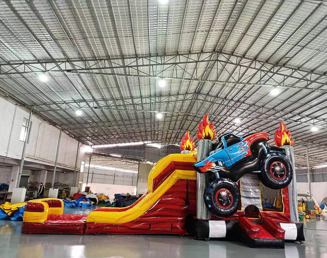 Wide-angle view of Monster Truck Dual Lane Commercial Inflatable Combo set up in a large indoor warehouse. The inflatable features a blue monster truck with flame decals, dual slides in red and yellow, and a splash pool area. Metal roofing, bright lighting, and various other inflatable products are visible in the background, demonstrating its size and commercial setting.
