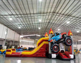 Wide-angle view of Monster Truck Dual Lane Commercial Inflatable Combo set up in a large indoor warehouse. The inflatable features a blue monster truck with flame decals, dual slides in red and yellow, and a splash pool area. Metal roofing, bright lighting, and various other inflatable products are visible in the background, demonstrating its size and commercial setting.