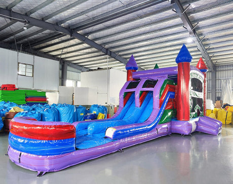 Wide-angle view of Multi Color Marble Splash & Save Dual Lane Inflatable Combo in a spacious warehouse. The inflatable displays vibrant purple, blue, and red colors with castle towers, dual slides, and a large splash pool. Various inflatable products, storage bags, and warehouse equipment are visible, demonstrating its size and commercial setting.