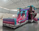 Three-quarter view of Pink Dino Dive Dual Lane Commercial Inflatable Combo set up in a warehouse. The structure features a pink and purple base with inflatable palm trees, dual slides, and large green dinosaur figures at the entrance. A colorful splash pool and various inflatable obstacles are visible. The image showcases the combo's size and detailed prehistoric theme in a commercial setting.