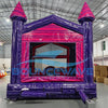 Front view of Purple Palace Commercial Grade Bounce House in a warehouse setting. The inflatable castle has purple walls, pink trim, and two tall towers with pink conical tops. A pink arched entrance and small slide are visible. The image shows the bounce house's size relative to the warehouse space, with cardboard boxes and other inflatables in the background.