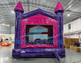 Front view of Purple Palace Commercial Grade Bounce House in a warehouse setting. The inflatable castle has purple walls, pink trim, and two tall towers with pink conical tops. A pink arched entrance and small slide are visible. The image shows the bounce house's size relative to the warehouse space, with cardboard boxes and other inflatables in the background.
