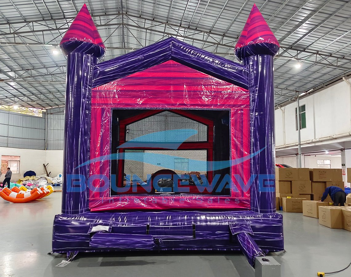 Front view of Purple Palace Commercial Grade Bounce House in a warehouse setting. The inflatable castle has purple walls, pink trim, and two tall towers with pink conical tops. A pink arched entrance and small slide are visible. The image shows the bounce house's size relative to the warehouse space, with cardboard boxes and other inflatables in the background.