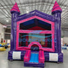 Wide-angle front view of Purple Palace Commercial Grade Bounce House in an indoor facility. The purple and pink inflatable features two tall towers, mesh windows, and a small front slide. Safety instructions are visible on the base. The warehouse setting shows metal roofing, fluorescent lighting, and various inflatable products and equipment in the background, demonstrating its commercial use context.