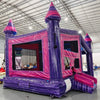 Three-quarter view of Purple Palace Commercial Grade Canopy Bounce House in an indoor facility. The purple inflatable structure has pink accents, four tall towers with conical tops, and a canopy roof. Mesh windows and a small slide are visible. The warehouse setting shows metal roofing, fluorescent lighting, and various inflatable products and equipment in the background, showcasing its size and commercial application.