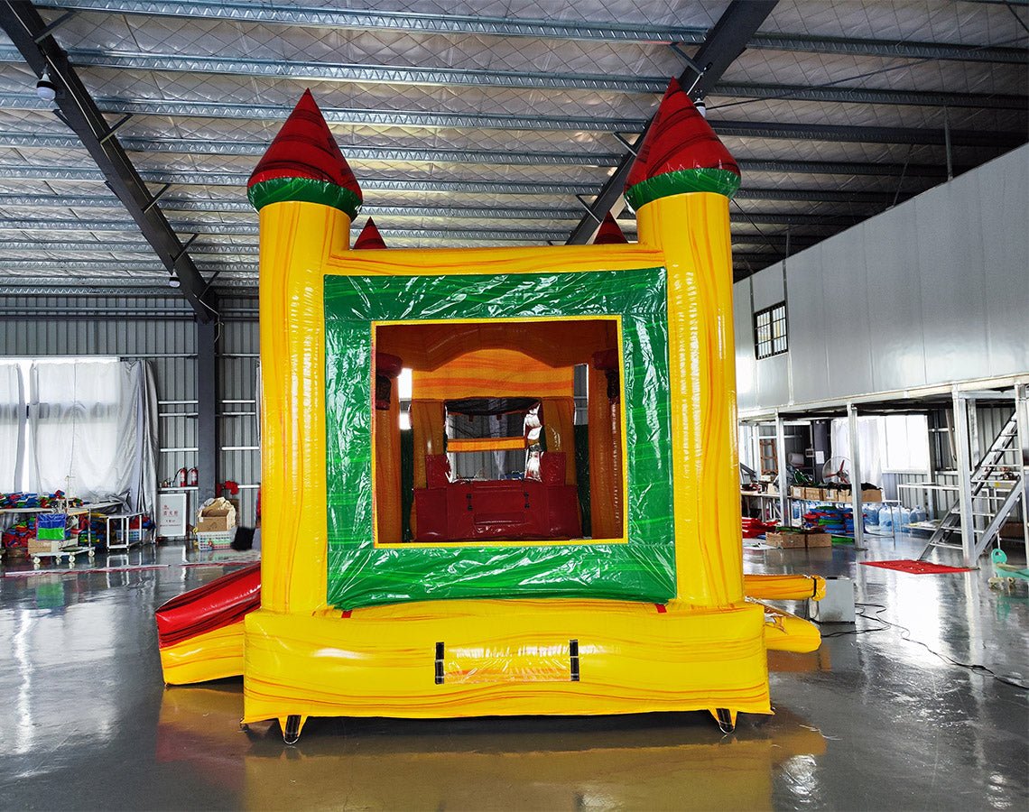 Front view of Rasta Splash Commercial Econo Inflatable Combo (Wet/Dry) set up in an indoor warehouse. Features yellow walls, red turrets, green frame, and a mesh-covered entrance. The inflatable includes a curved slide and splash pool area. Metal roofing and other warehouse equipment visible in the background.