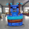 Large inflatable tropical-themed combo featuring a bounce house with palm tree decorations, attached slide, and splash pool. The structure has vibrant blue and turquoise colors with brown palm tree trunks and a large inflatable sunglasses decoration on top. Set up in an indoor warehouse with metal roofing visible.