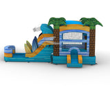 Large inflatable beach-themed combo featuring a bounce house with palm tree decorations, attached wave-shaped slide, and play area. The structure has vibrant blue, yellow, and green colors with surfboard designs. Four inflatable palm trees top the bounce house. Mesh windows allow visibility inside. Set against a white background.