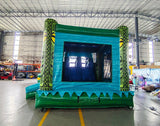 Wide-angle view of Tiki Shot Curved Dual Lane Commercial Inflatable Combo (Wet/Dry) in a spacious warehouse. Features a large tiki-faced tower with green pillars and yellow eyes, dual curved blue slides, and a circular splash pool with yellow and green accents. The inflatable structure has vibrant turquoise, green, and yellow colors. Warehouse interior visible with metal roofing, shelving units, and other equipment, providing scale and context for the inflatable's size.