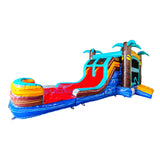 Side view of Tropic Shock Dual Lane Commercial Inflatable Combo featuring a tropical theme with turquoise palm trees. The structure has dual red slides emerging from a turquoise frame, brown tree trunk pillars, and a colorful base with blue, yellow, and red sections. A large circular splash pool area is visible at the front. Set against a white background, showcasing its vibrant design for commercial wet/dry use.