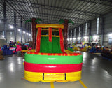 Front view of Tropical Inferno Dual Lane Commercial Inflatable Combo showing dual red slides descending into a green and yellow splash pool. The inflatable features brown palm tree trunks with green leaves, and a yellow top banner. Various other inflatable products and warehouse equipment are visible in the background, showcasing its commercial setting.