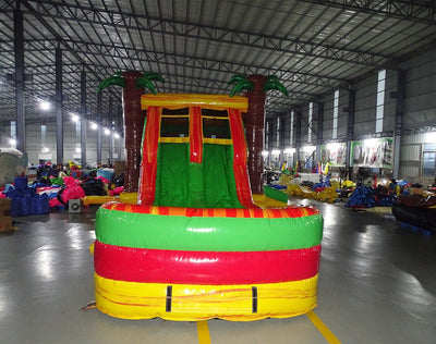Front view of Tropical Inferno Dual Lane Commercial Inflatable Combo showing dual red slides descending into a green and yellow splash pool. The inflatable features brown palm tree trunks with green leaves, and a yellow top banner. Various other inflatable products and warehouse equipment are visible in the background, showcasing its commercial setting.
