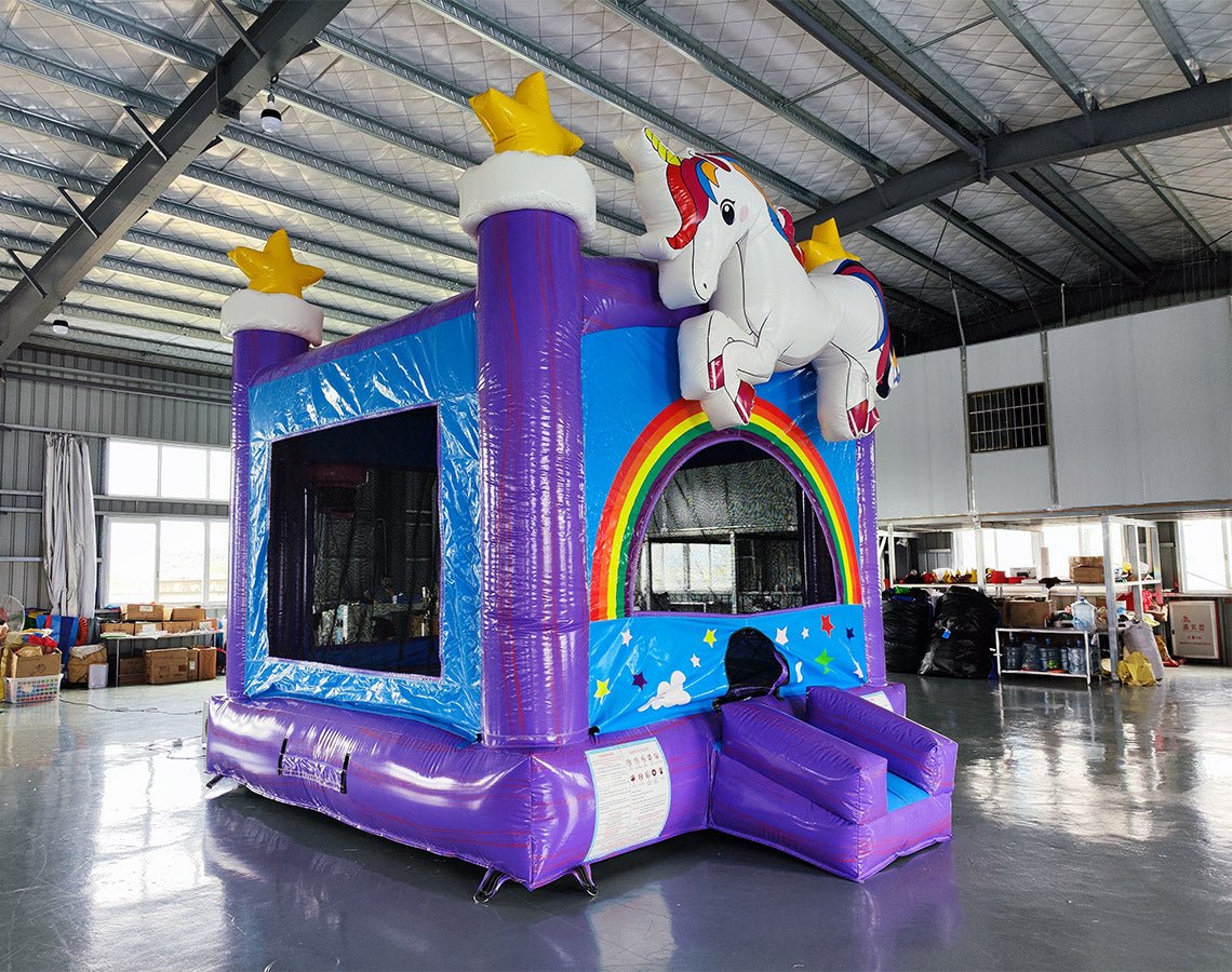 Wide-angle view of Unicorn Commercial Grade Bounce House in a large indoor warehouse. Purple inflatable structure with blue panels, rainbow arch entrance, and white unicorn figure on top. Yellow stars adorn purple towers. Visible warehouse features include metal roofing, windows, and various equipment, demonstrating the bounce house's size and commercial setting.