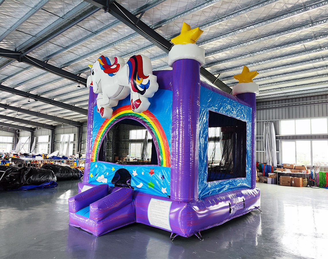 Three-quarter view of Unicorn Commercial Grade Bounce House in warehouse setting. Purple inflatable with blue panels, featuring white unicorn atop rainbow arch. Yellow stars on purple towers. Small blue slide at entrance. Warehouse ceiling, windows, and various equipment visible, showcasing the bounce house's size and commercial application.