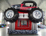 side view of UTV Trail Boss Commercial Grade Bounce House set up in a large indoor warehouse. The inflatable features a realistic off-road vehicle design with large black tires, a red and black body, and a roll cage. The bounce area is visible beneath the vehicle. Warehouse interior with metal roofing and various inflatable products visible in the background.